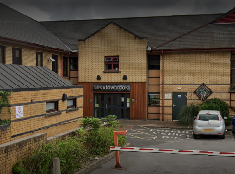 The image shows Meadowbrook acute mental health unit in Salford. The front of the building can be see with the Meadowbrook sign above it. There is also a small section of car park in the frame.